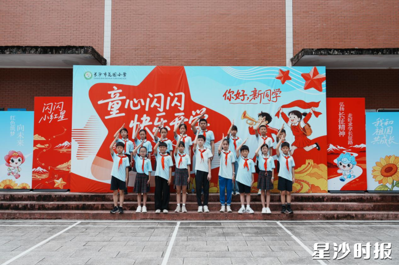 长沙市花园小学开学现场，少先队员们在红色主题背景板前整齐列队敬礼，红色教育浸润开学第一课。